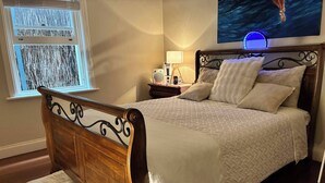 Un lit bien fait, avec une tête de lit en bois, une table de chevet avec une lampe et une fenêtre équipée de stores.