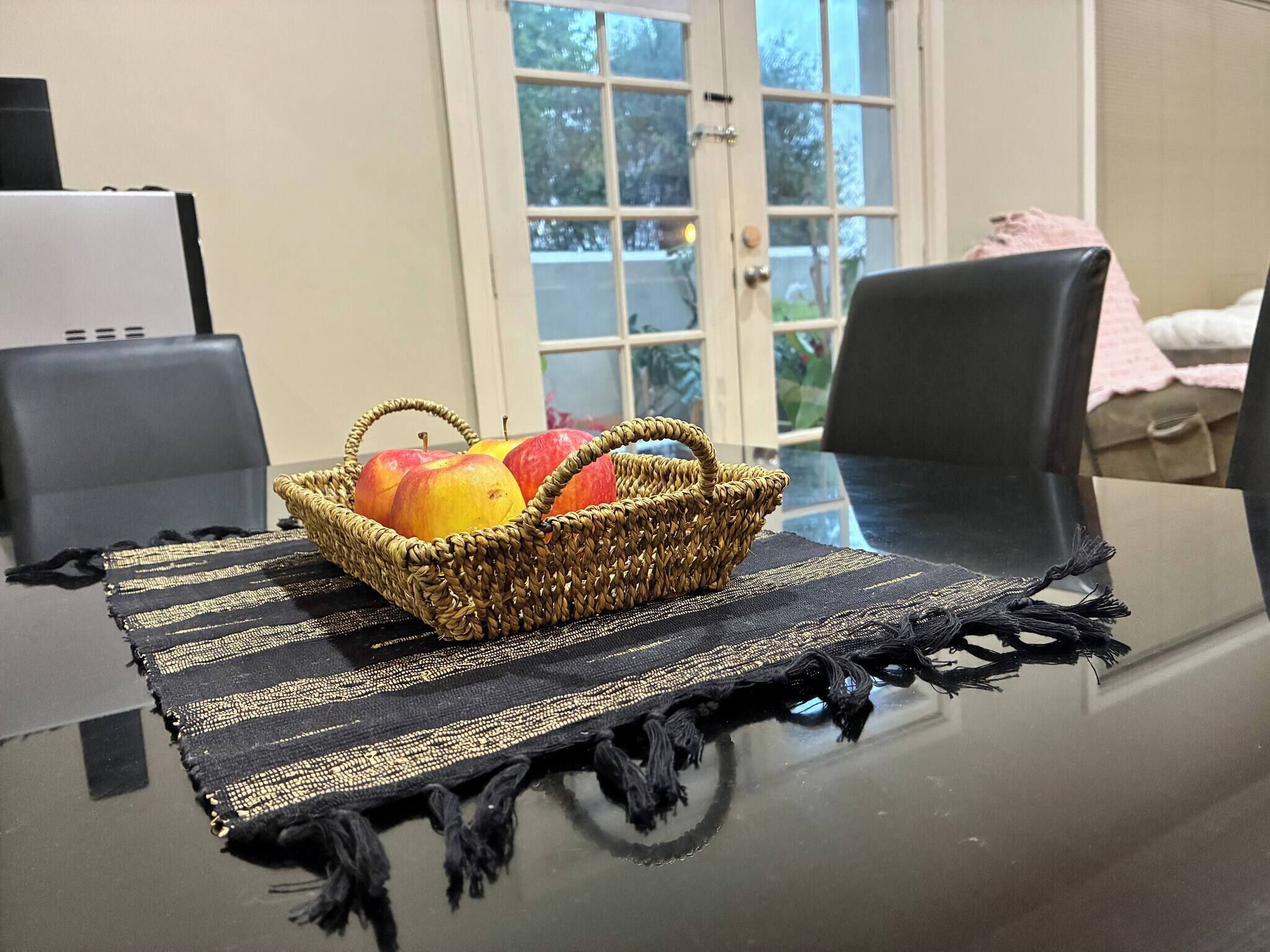 A table with a woven basket containing apples, a black and gold woven placemat, and a glass table.