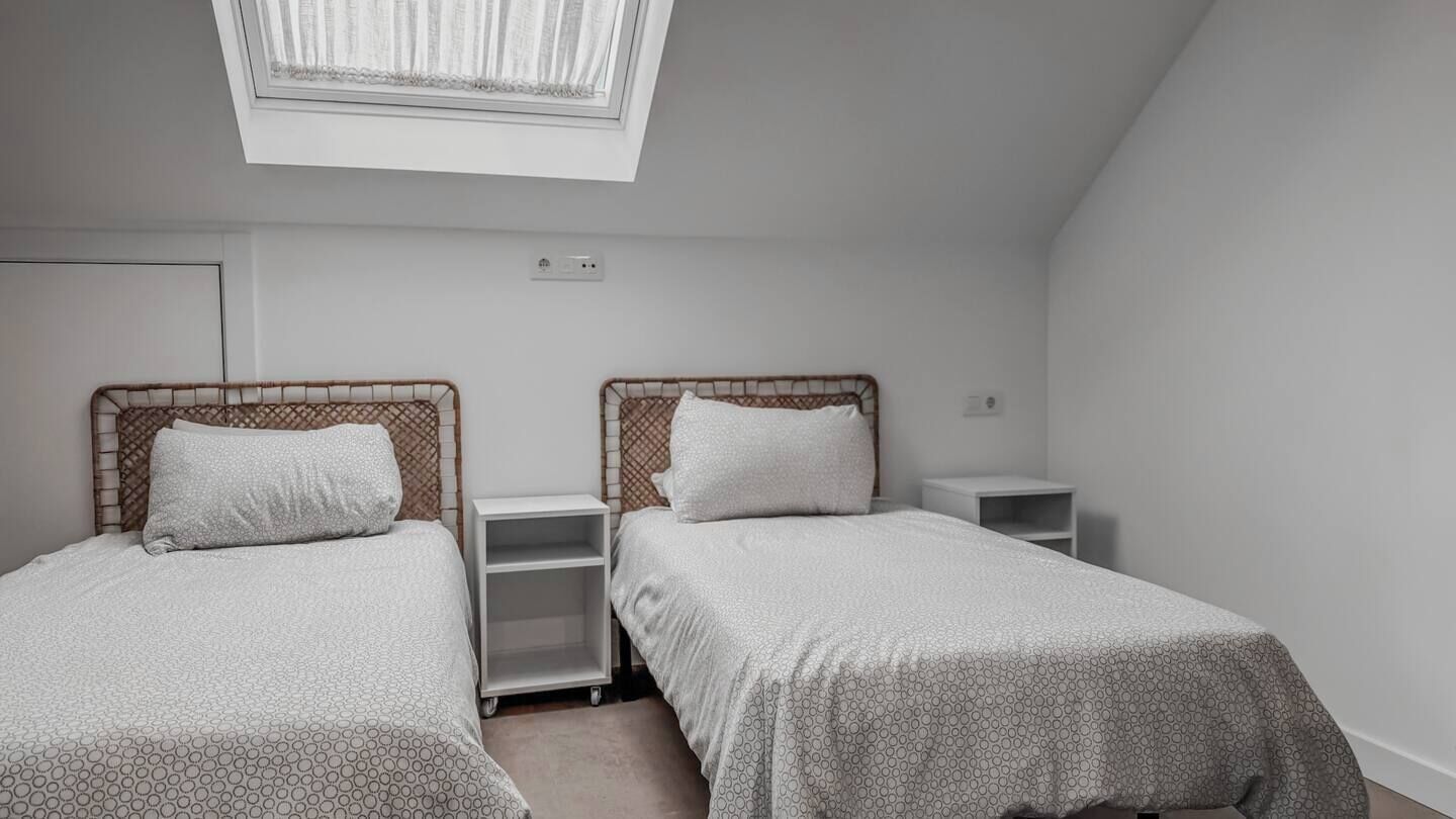 Un dormitorio con dos camas individuales, una mesita de noche pequeña y blanca, un estante blanco y un tragaluz.