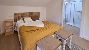 Een slaapkamer met een bed, een bankje, een plank en een douchecel zichtbaar door een geopende deur.