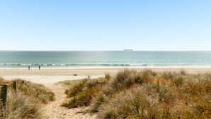 A beach with a sandy path leading to the water, a ship in the distance, and grassy dunes. - Cabbage Tree Cottage (Tauranga)