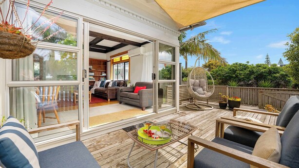 A spacious outdoor deck with a covered seating area, a dining table, and a hanging plant. - Cabbage Tree Cottage (Tauranga)