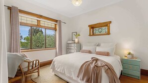 A bedroom with a large window, a bed with white bedding, a bedside table with a lamp, and a small plant. - Cabbage Tree Cottage (Tauranga)