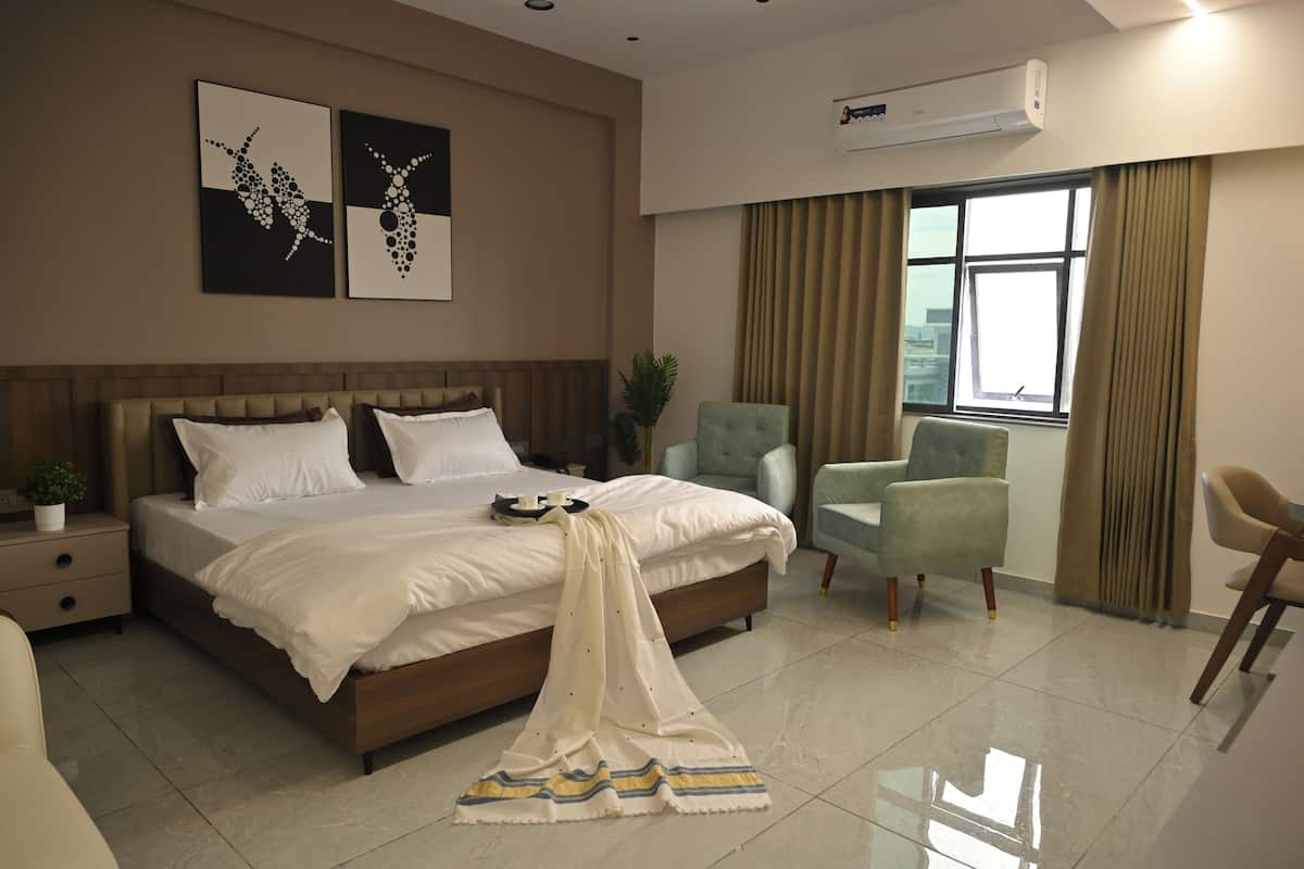 Una habitación de hotel con una cama, dos sillones, una mesita de noche, una planta en maceta y obras de arte en la pared.