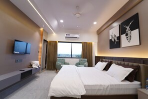 A modern hotel room with a large bed, a flat-screen TV, a seating area with two chairs, and artwork on the walls. - Plumeria Hotel  (Bhuj)