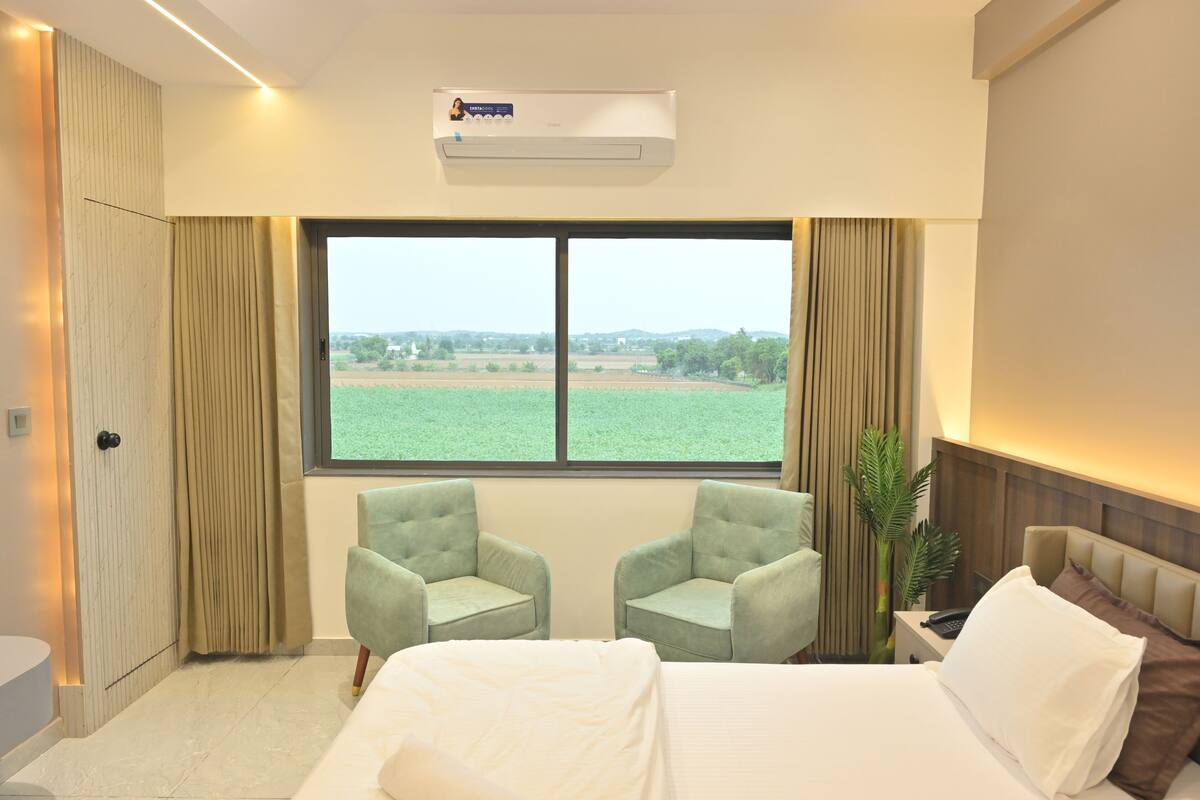 Una habitación de hotel con un ventanal grande, dos sillones, una cama y un equipo de aire acondicionado.
