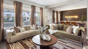 A modern living room with a sectional sofa, a round coffee table, and a dining area with a neon sign. - The Nowland Reserve |Sauna, Arcade, Sleeps 14 (Indianapolis)