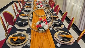 A long dining table set for a formal event, with chairs on both sides and a stage in the background.