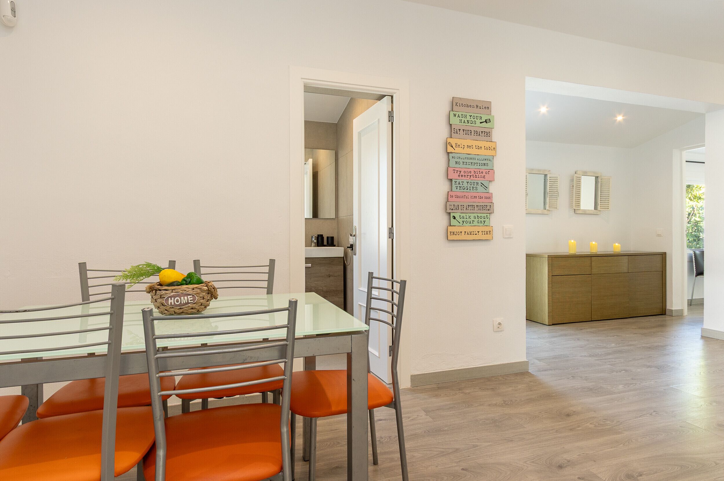 A dining area with a glass table, orange chairs, and a fruit basket labeled 'HOME'.