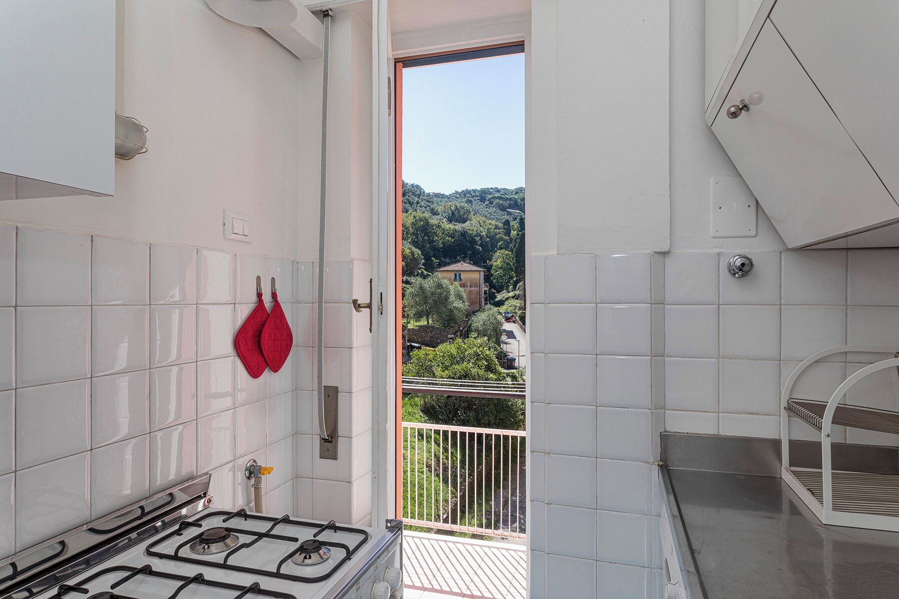 A kitchen with white tiles, a gas stove, and a view of a green landscape through an open door.