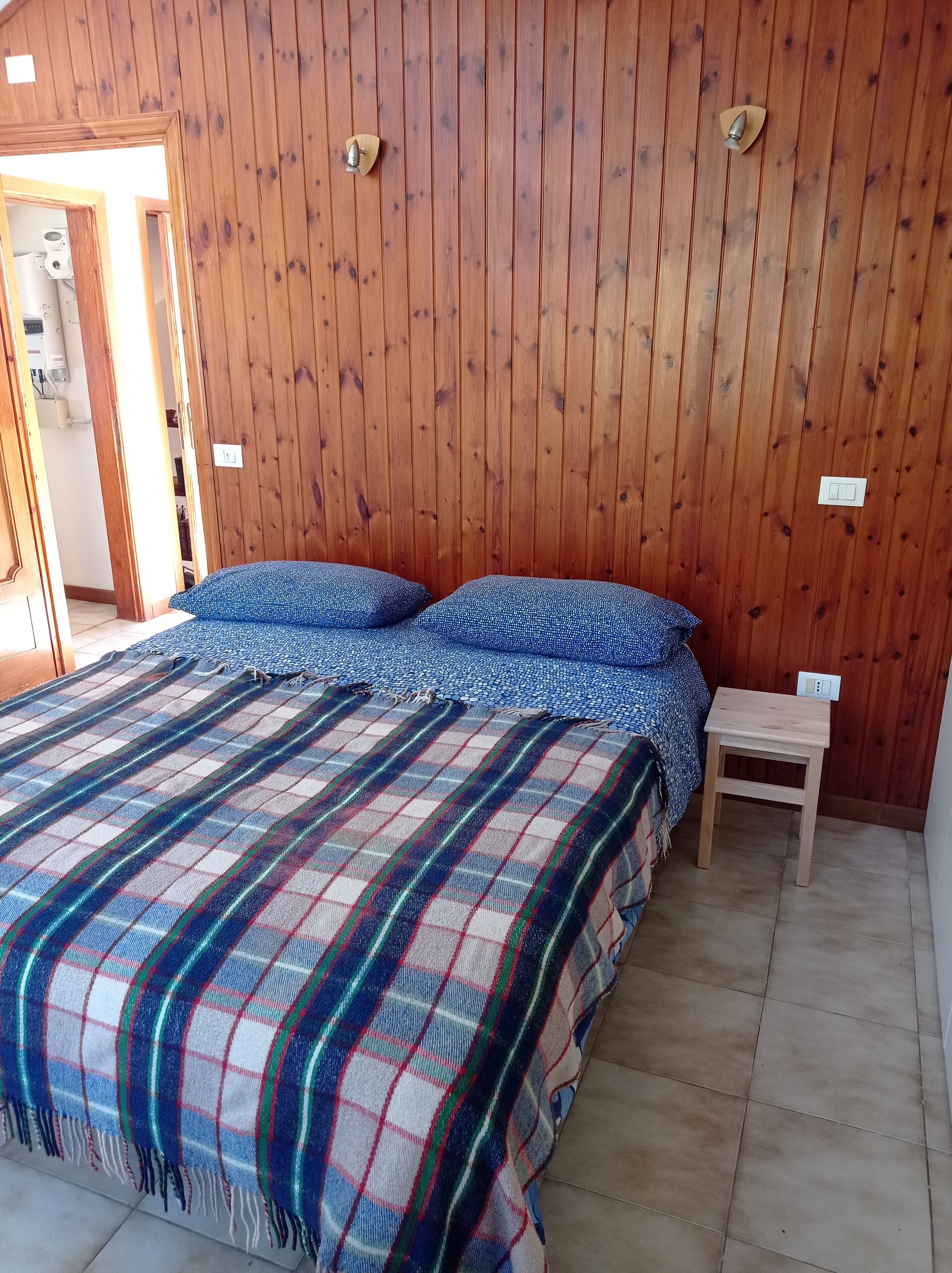Ein Zimmer mit Holzwänden, einem Bett, einem kleinen Holzschemel und einer Tür zu einem anderen Raum.