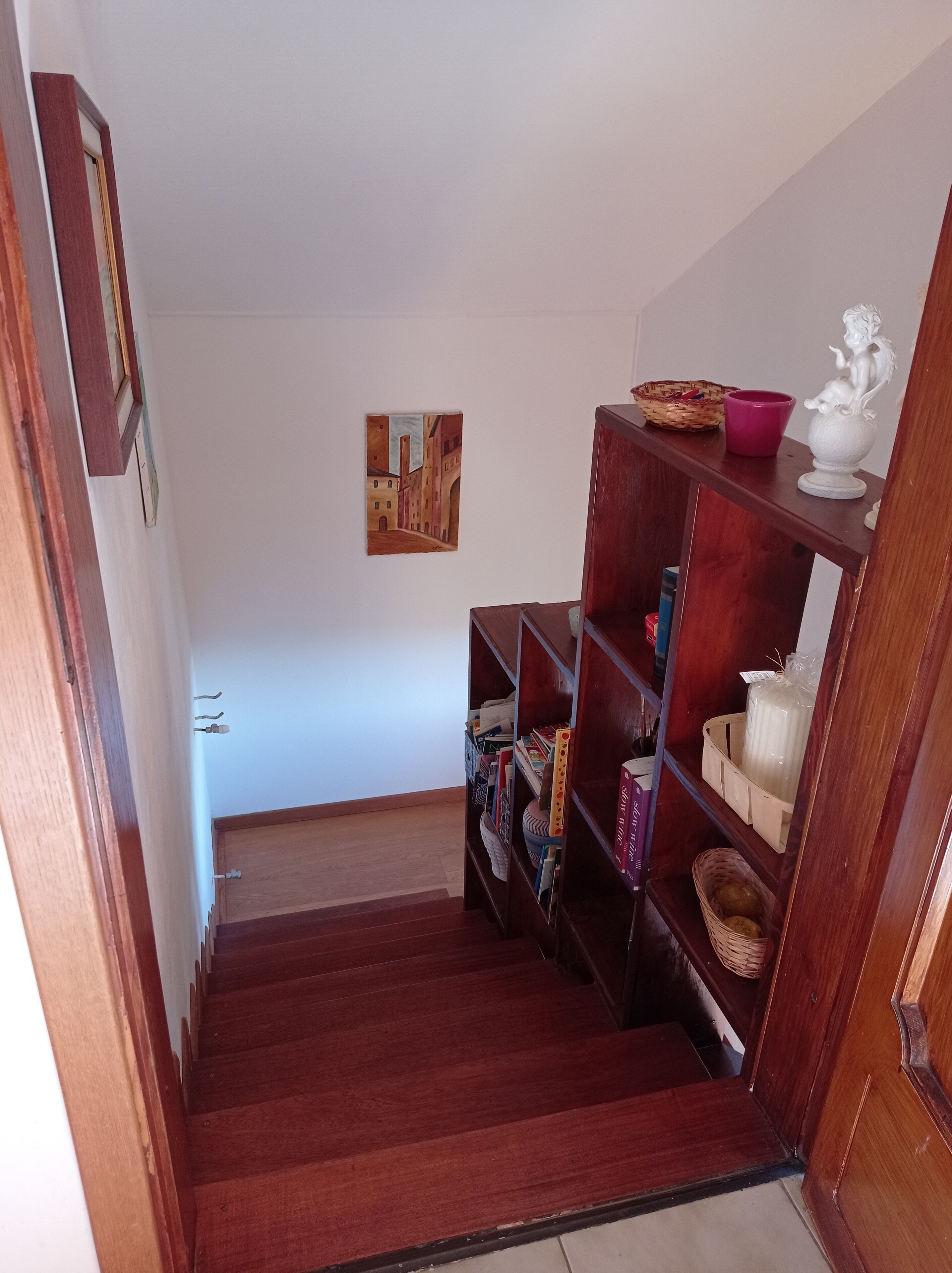 Ein Flur mit einer Holztreppe, die zu einem Zimmer führt. Es gibt ein Bücherregal mit Büchern und Dekoration, ein kleines Gemälde an der Wand und einen Korb auf der obersten Ablagefläche.