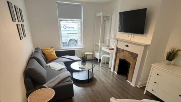 A modern living room with a sofa, coffee table, and white armchair. There is a fireplace, a television mounted on the wall, and a window with a view of a car outside. - Corner Stone House Apartment 2 (Great Yarmouth)