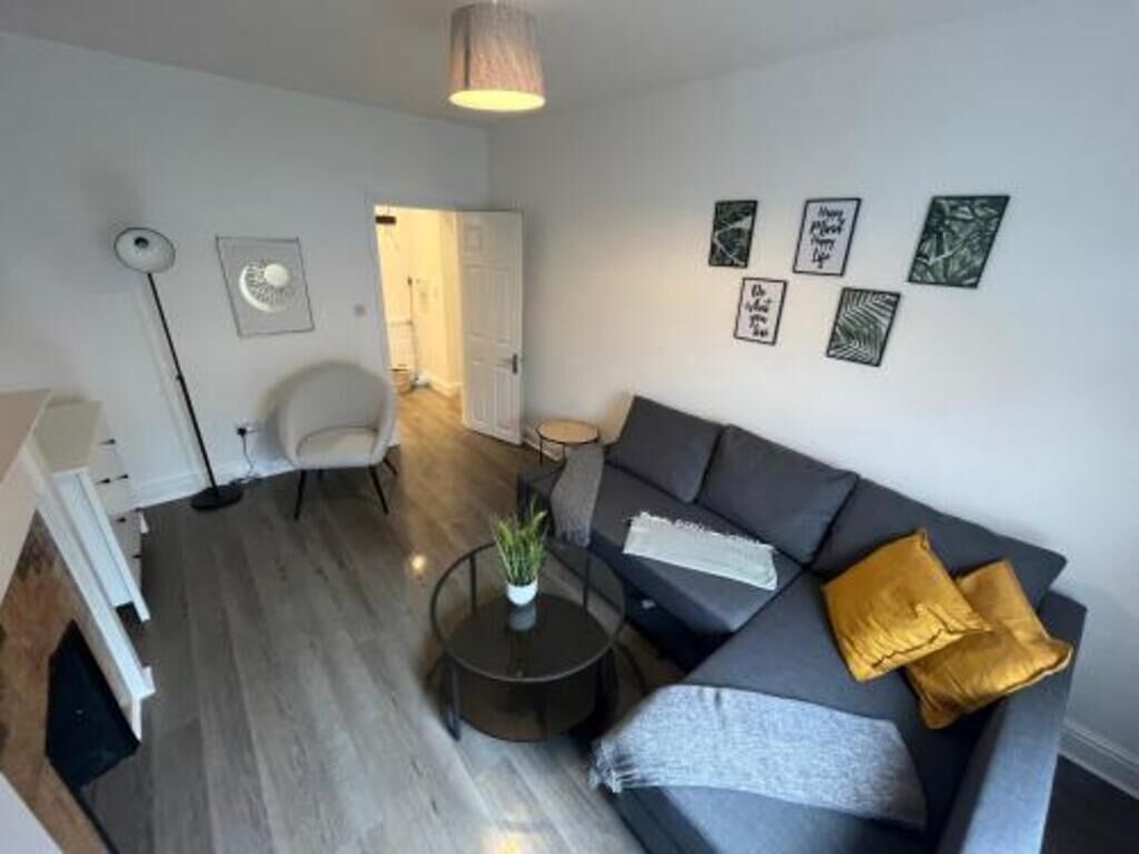 Un salon moderne avec un canapé gris, une petite table basse ronde et un fauteuil blanc. Des tableaux encadrés ornent le mur et une lampe est suspendue au plafond.
