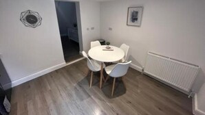 A modern dining area with a round table and white chairs, a white radiator, and a geometric wall decoration. - Corner Stone House Apartment 2 (Great Yarmouth)