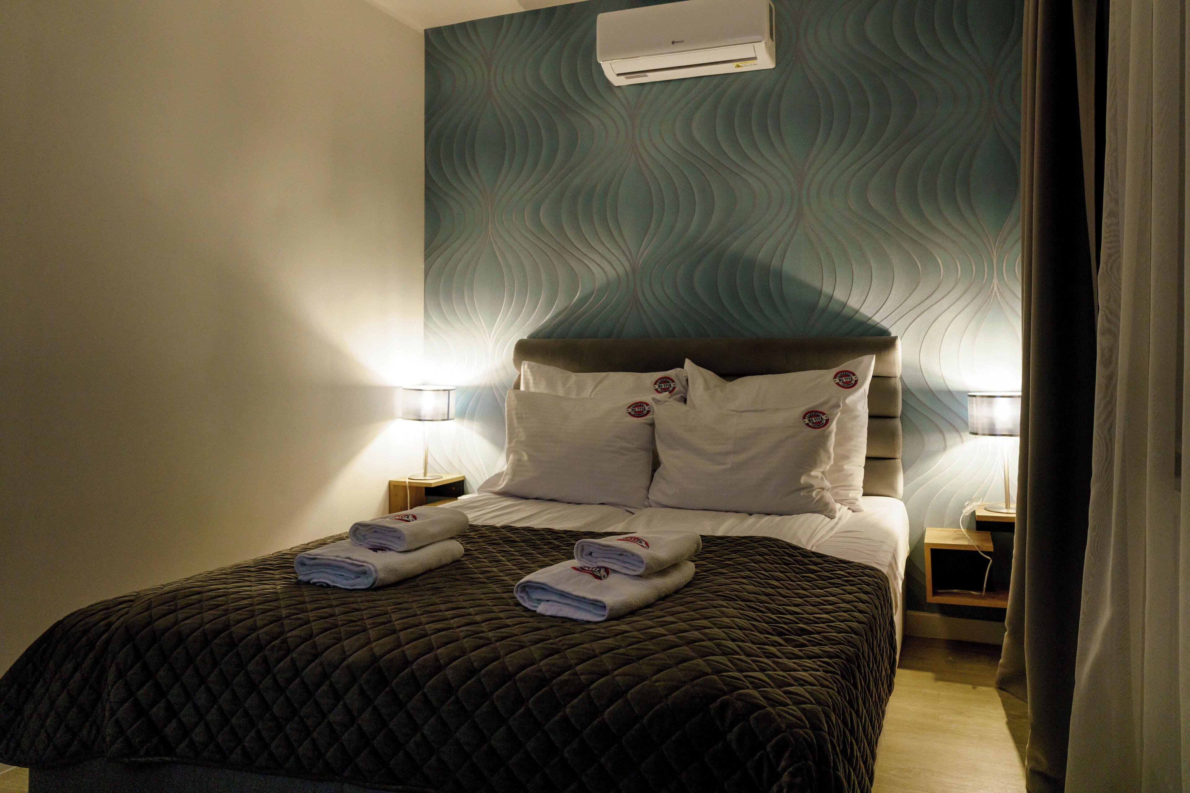 A neatly made bed with a quilted cover, two pillows, and folded towels on the bed. There is a wall-mounted air conditioner above the bed, and two bedside lamps on either side.