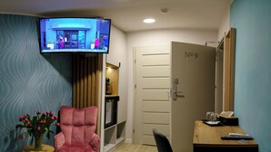 A hotel room with a television displaying news, a pink armchair, a wooden sideboard, a desk with a computer, and a door labeled "No 9". - Kamienica no111a (Warsaw)