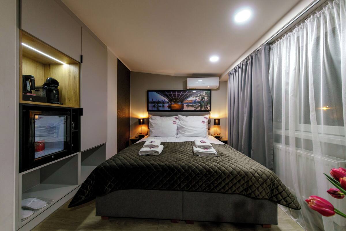 Una camera d'albergo moderna con un letto grande, una TV a schermo piatto fissata al muro, una macchina del caffè e una finestra con tende.