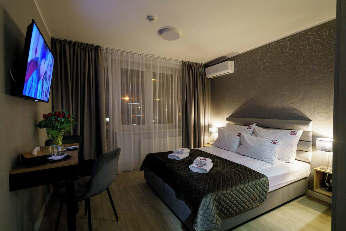 Una camera d'albergo con un letto, una scrivania con televisore, una sedia e una finestra con tende.