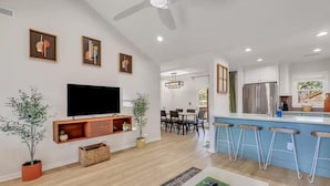 Ein modernes Wohnzimmer mit Flachbildfernseher, Holzböden, Blumentöpfen, einem Essbereich mit Tisch und Stühlen sowie einer Küche mit Edelstahlkühlschrank und blauen Barhocker.