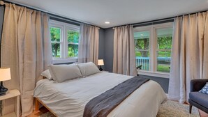 A bedroom with a bed, two bedside tables with lamps, a chair, and large windows with curtains. - Marvelous Manning: 4-Bdr + Studio + Gameroom (Shad Moss village)