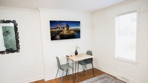 A dining area with a table and chairs, a framed picture on the wall, and a window with blinds. - The Palmetto Place - 2br Modern Unit on Remount (North Charleston)