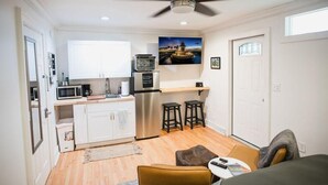 A compact kitchen with white cabinets, a microwave, a refrigerator, and a ceiling fan. There is a small seating area with a table and a chair. - The Grove Studio - Comfort in Style on Remount (North Charleston)
