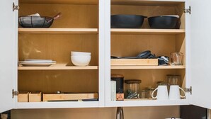 A kitchen with open cabinets, containing bowls, plates, a coffee maker, and utensils. - The Grove Studio - Comfort in Style on Remount (North Charleston)