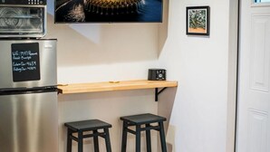 A kitchen with a stainless steel refrigerator, a wooden countertop, two black stools, a wall-mounted TV, and a framed picture. - The Grove Studio - Comfort in Style on Remount (North Charleston)