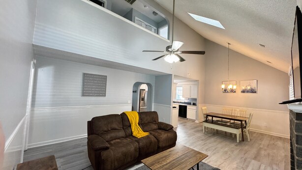 A spacious living room with a brown leather sofa, a wooden coffee table, and a ceiling fan. A dining area with a wooden table and chairs is visible in the background. - Bright & Roomy 4BR/5-Bed Home Just Off Dorchester (North Charleston)