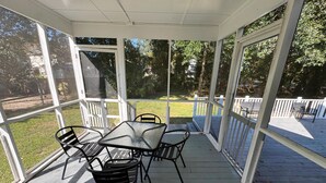 A covered outdoor patio with a table and chairs, surrounded by glass doors and windows, overlooking a grassy yard with a wooden deck. - Bright & Roomy 4BR/5-Bed Home Just Off Dorchester (North Charleston)