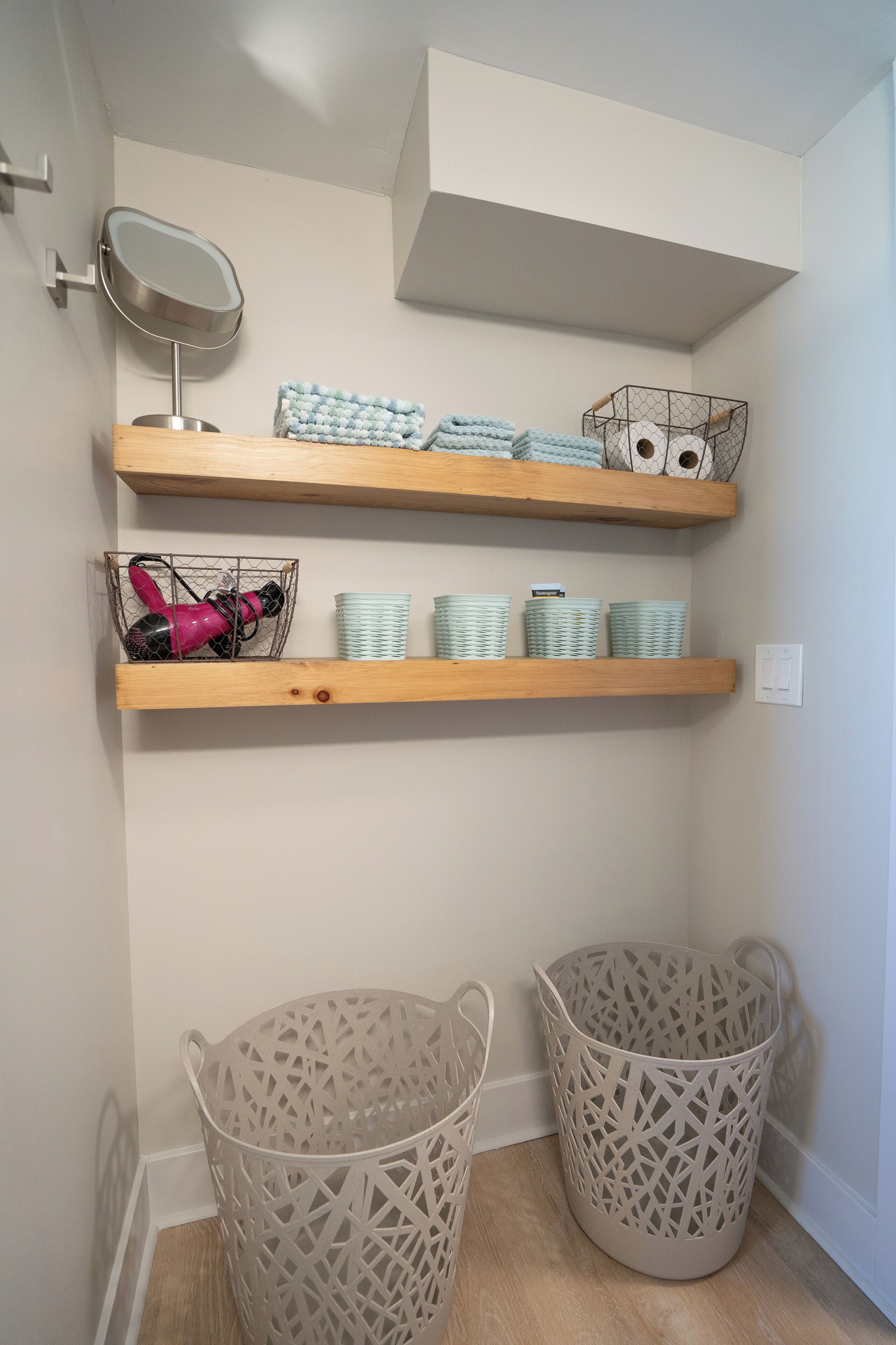 A corner of a room with two beige woven baskets, a mirror, and shelves with folded towels and stacked plates.