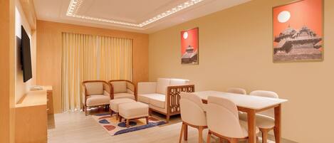 A modern hotel room with a wooden desk, a sofa, a dining table with chairs, and a TV.