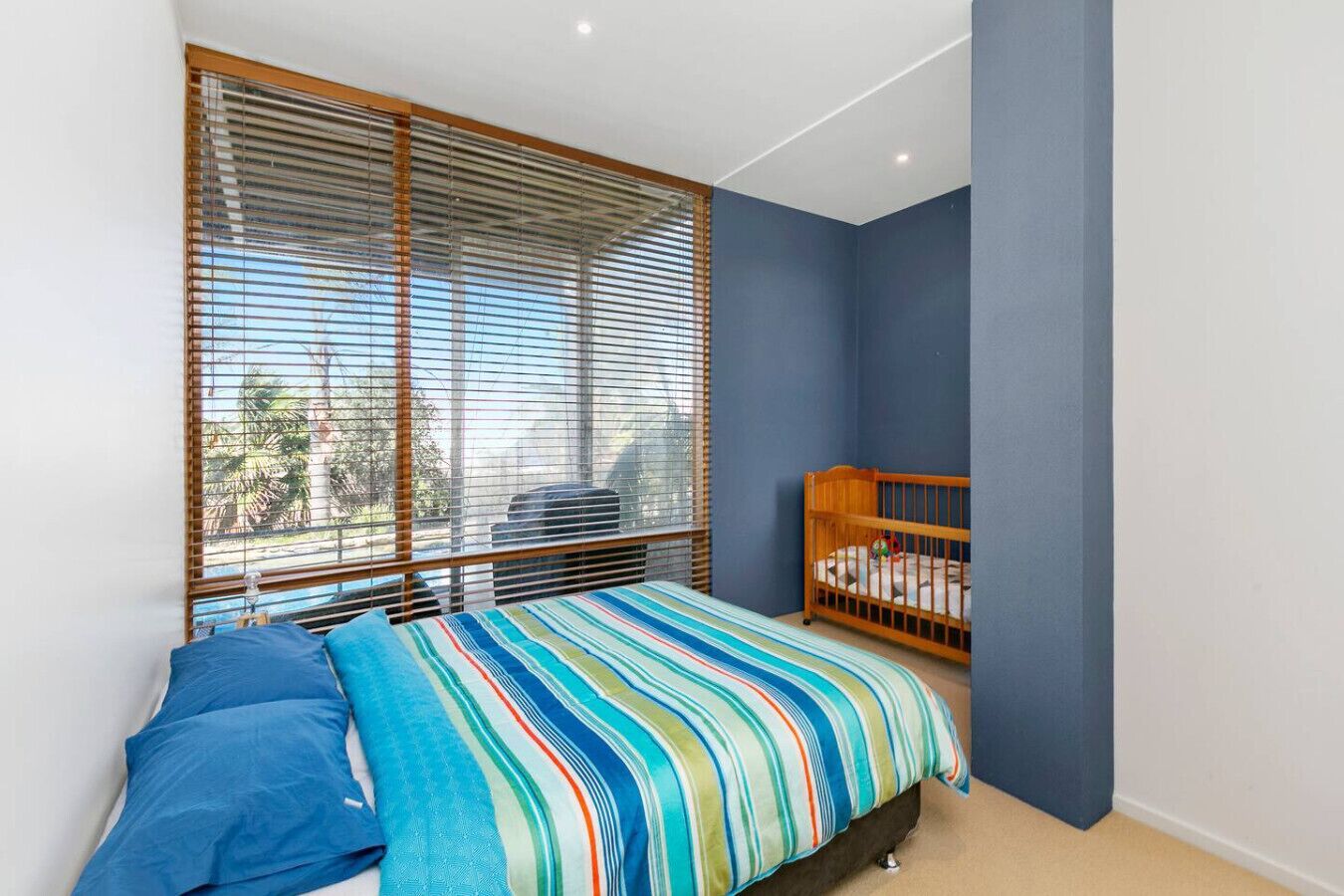 A bedroom with a bed, a crib, and large windows with blinds.