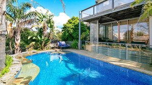 En swimmingpool med krystalklart blåt vand, omgivet af tropisk vegetation og en moderne bygning med store vinduer.