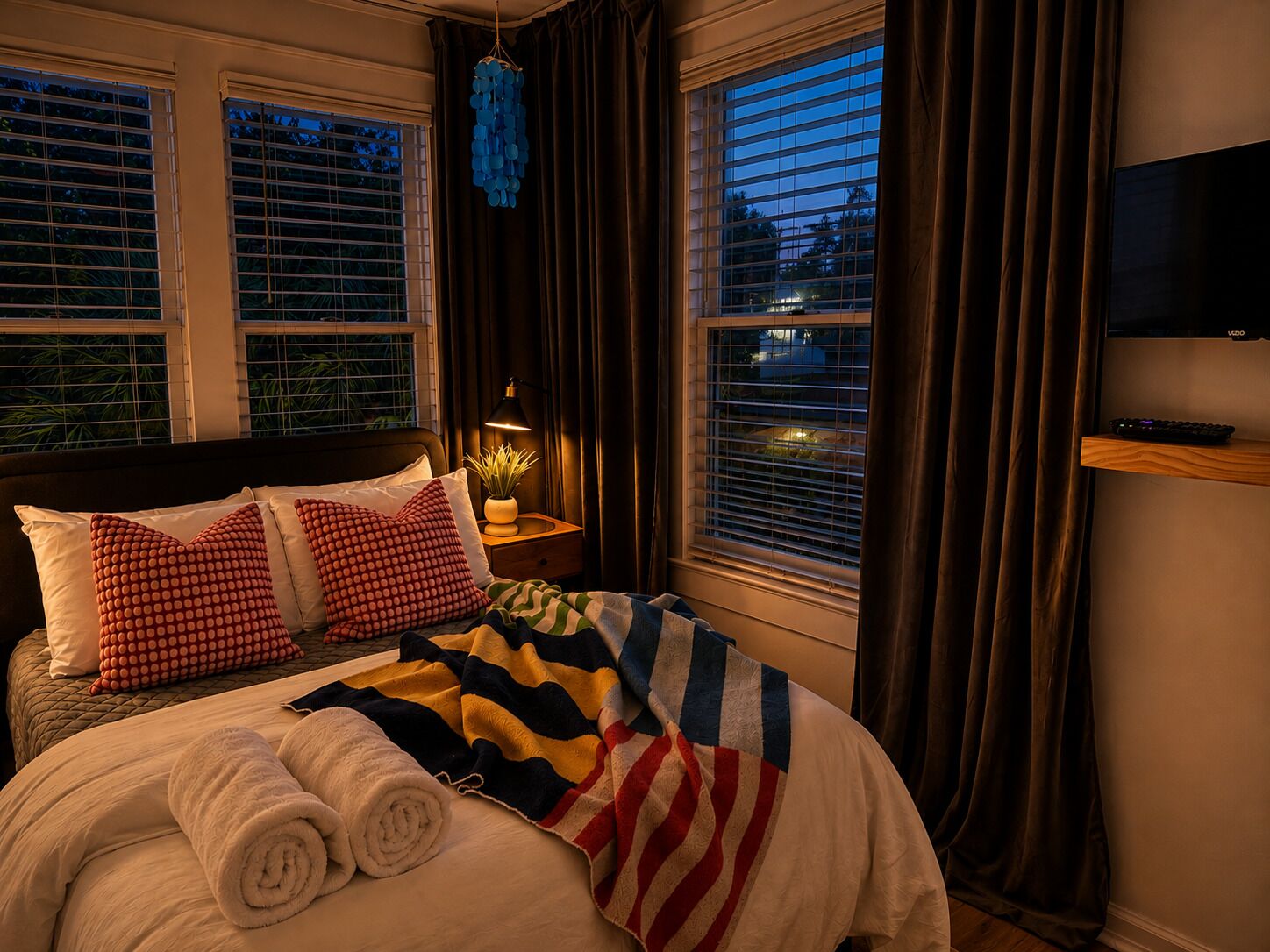Een hotelkamer met een bed, twee kussens, een nachtkastje met een lamp, een televisie aan de muur en ramen met gordijnen.