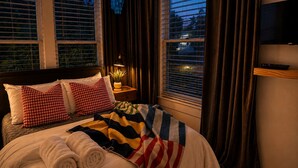 A hotel room with a bed, two pillows, a bedside table with a lamp, a TV on the wall, and windows with curtains.
