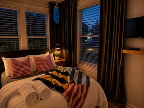 Una camera d'albergo con un letto, due cuscini, un comodino con una lampada, una TV a parete e finestre con tende.