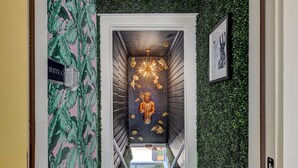 A narrow hallway with a tropical leaf-patterned wall, a chandelier, and a framed picture on the right.