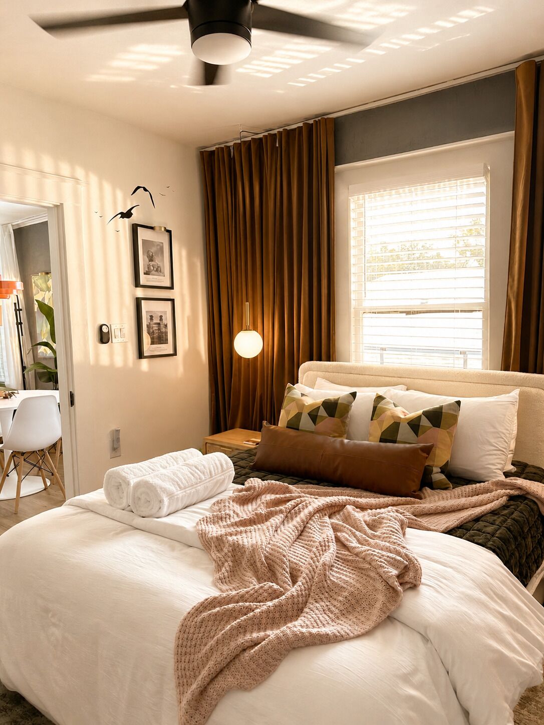 A hotel room with a bed, a bedside table, a lamp, a window with curtains, and a ceiling fan.