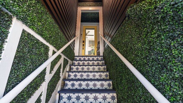 A staircase with a patterned floor, surrounded by green foliage walls, leading to a door with a light fixture above.