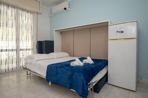 A bedroom with a bed, a blue blanket, a white refrigerator, and a window with curtains. - Apartment  Turchese (Pietra Ligure)