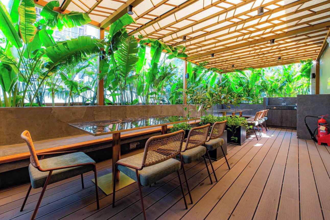 An outdoor seating area with wooden tables and chairs, surrounded by lush tropical plants.