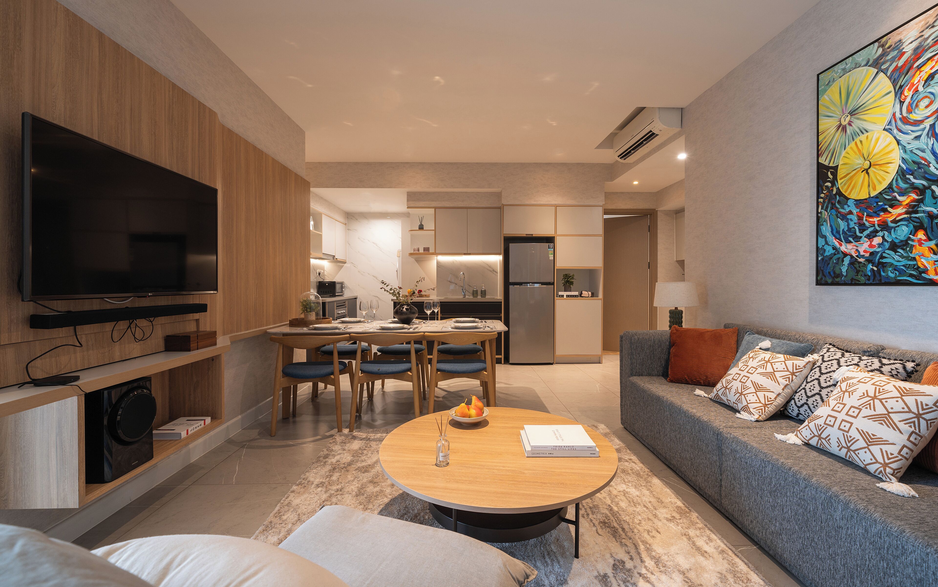 A modern living room with a flat-screen TV, a dining area with a wooden table and chairs, a grey sofa with patterned cushions, and a kitchen in the background.
