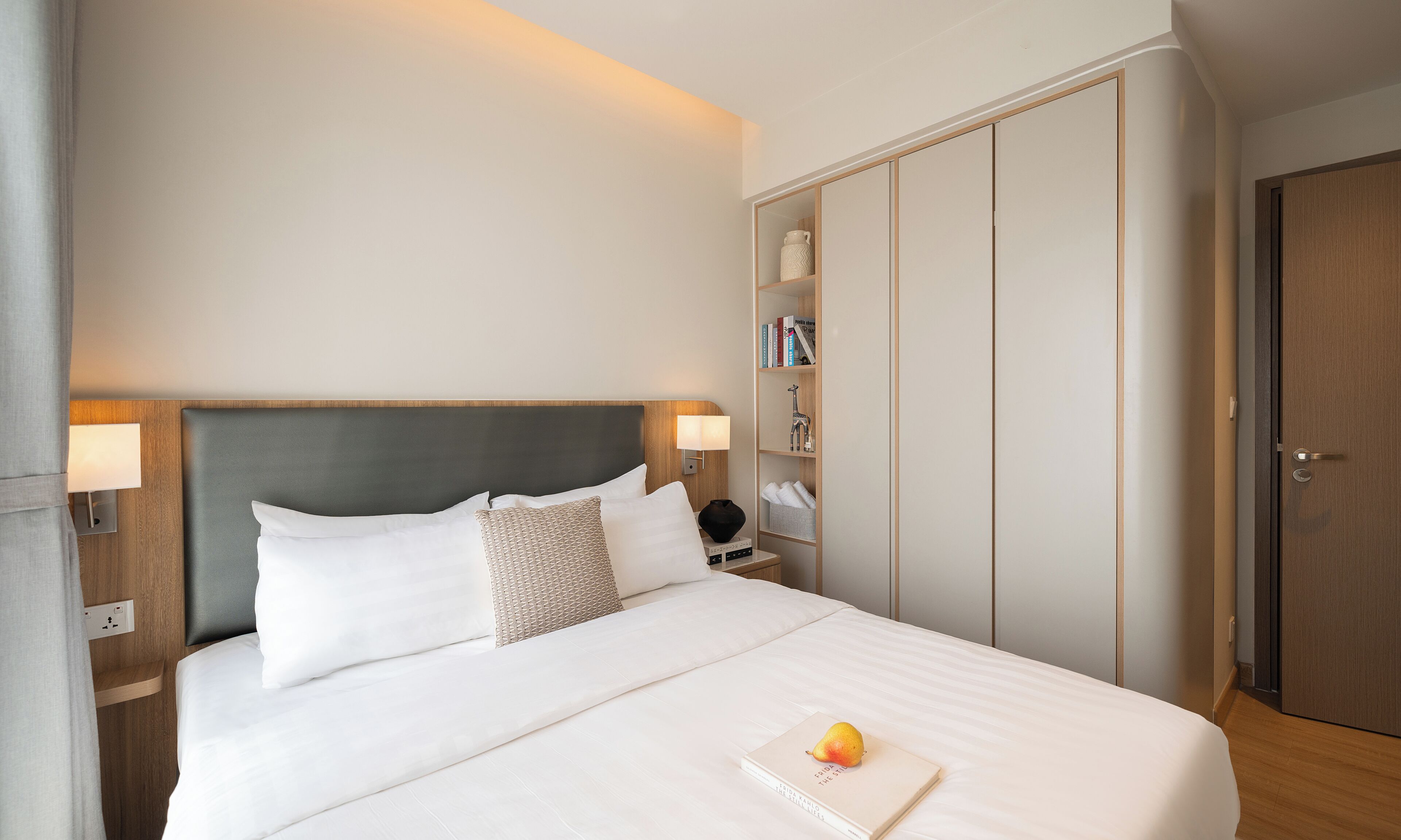 Un lit bien fait, avec du linge de lit blanc, une lampe de chevet et un livre sur lequel repose un fruit. On aperçoit une armoire en bois avec un miroir et une porte sur le côté droit.