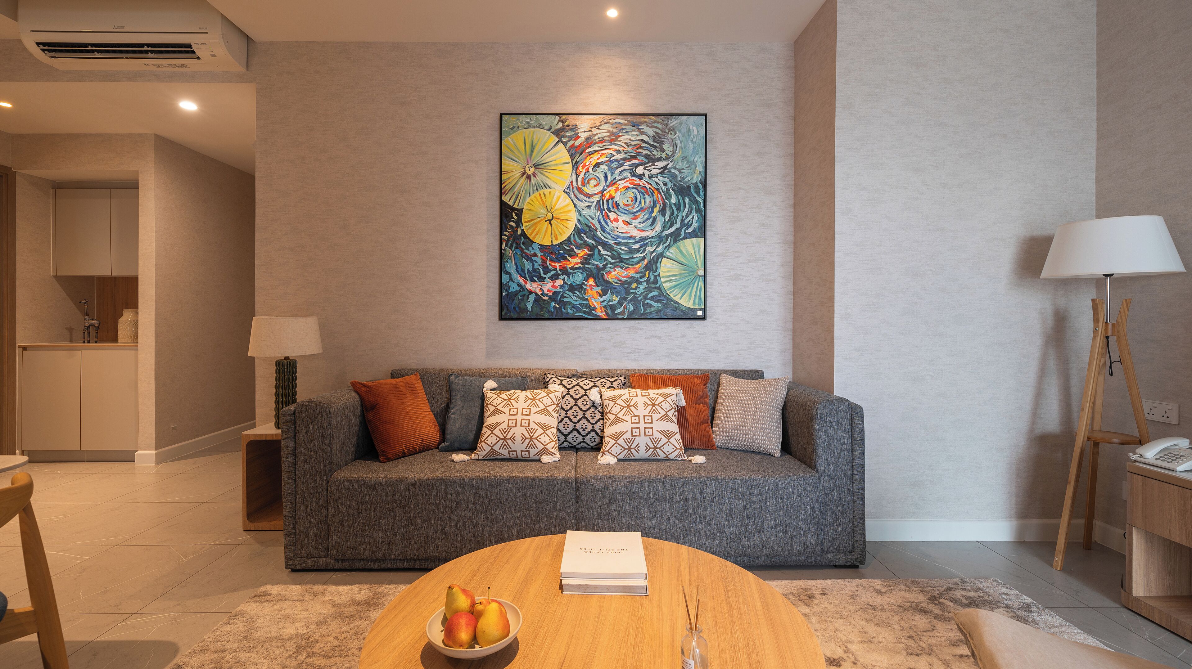 A modern living room with a grey sofa, a round wooden coffee table with a book and fruit bowl, and a colorful abstract painting above the sofa.