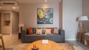 A modern living room with a grey sofa, a round wooden coffee table with a book and fruit bowl, and a colorful abstract painting above the sofa.