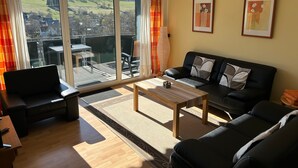 A living room with a sofa, a coffee table, and a view of a green landscape through large windows.