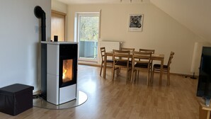 A modern living room with a wood-burning stove, a dining area with wooden chairs, and a large window with a view of trees.