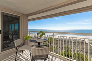 Varanda com vista para a praia, móveis de vime e uma mesa com uma planta e copos de vinho.